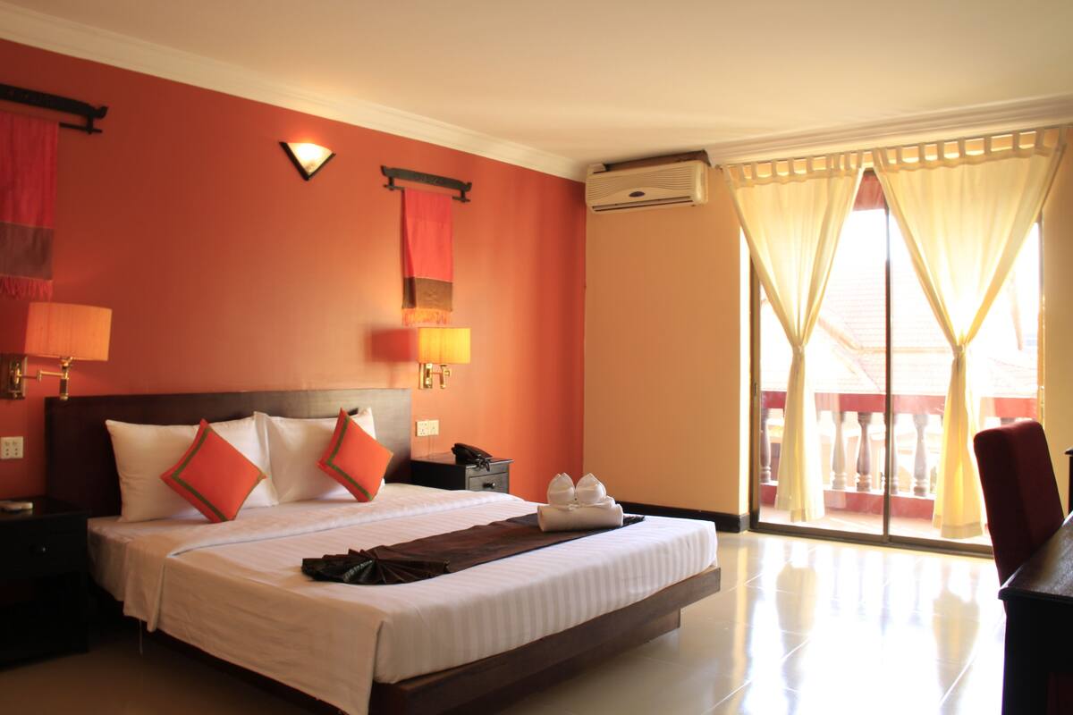 Habitación de hotel con una cama grande, una mesita de noche, una lámpara, aire acondicionado instalado en la pared y vistas al exterior a través de una puerta corrediza de vidrio.