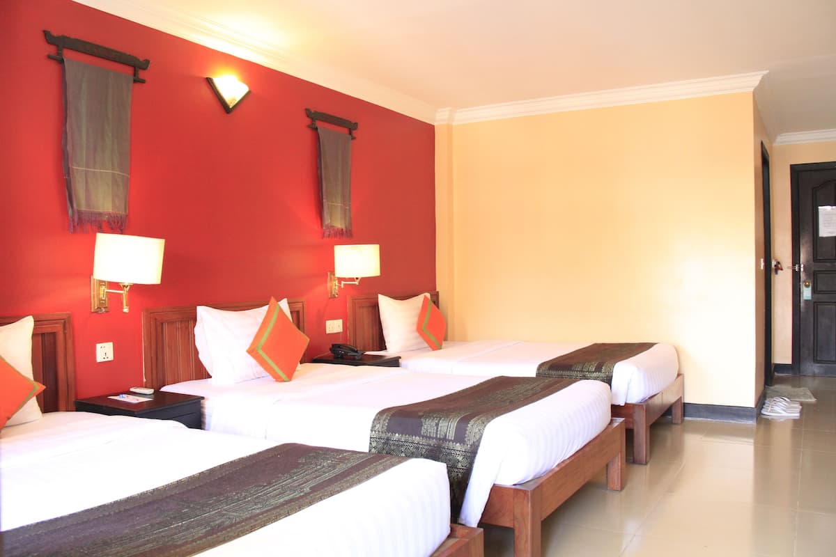 Habitación de hotel con tres camas individuales, cada una con cabecero de madera y ropa de cama blanca. Hay dos mesitas de noche con lámparas, y las paredes están pintadas de rojo y amarillo.