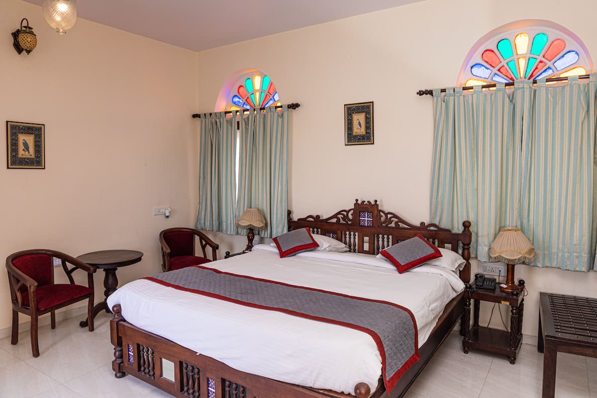 Une chambre d’hôtel avec un grand lit, deux chaises, une petite table, une table de chevet et une fenêtre avec des rideaux.
