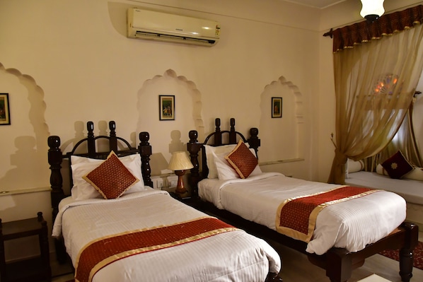 A hotel room with two beds, each with a wooden headboard, white bedding, and red accents. There is a wall-mounted air conditioner and a lamp on a bedside table.