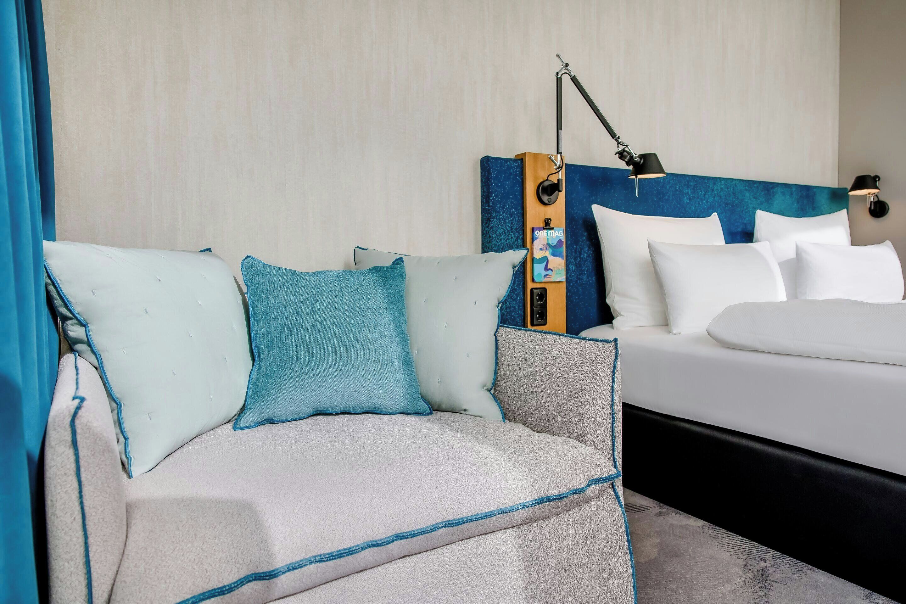 A hotel room with a sofa and a bed, a headboard with a clock, and a wall-mounted lamp.