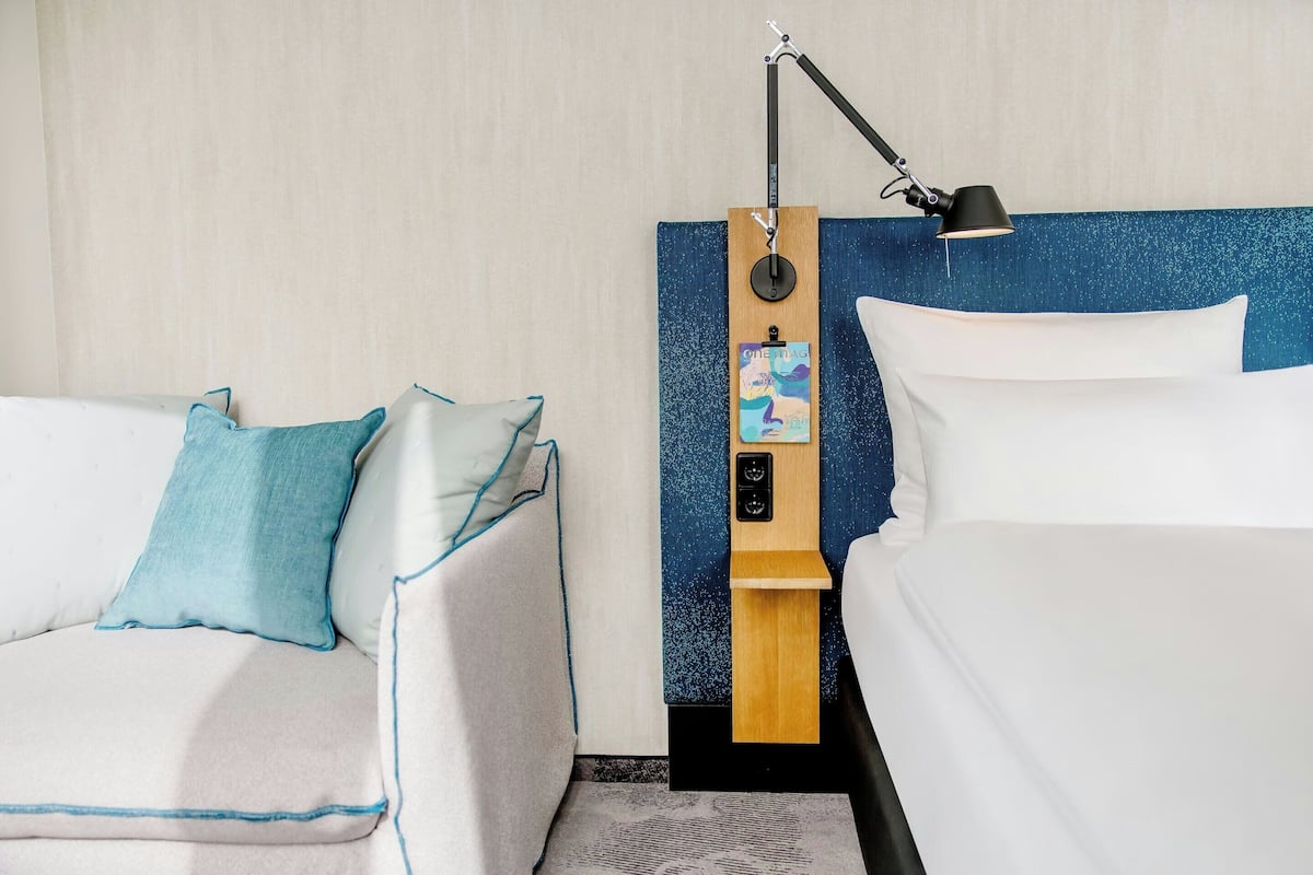 A hotel room with a bed, a sofa, a nightstand with a lamp, and a wall-mounted control panel.