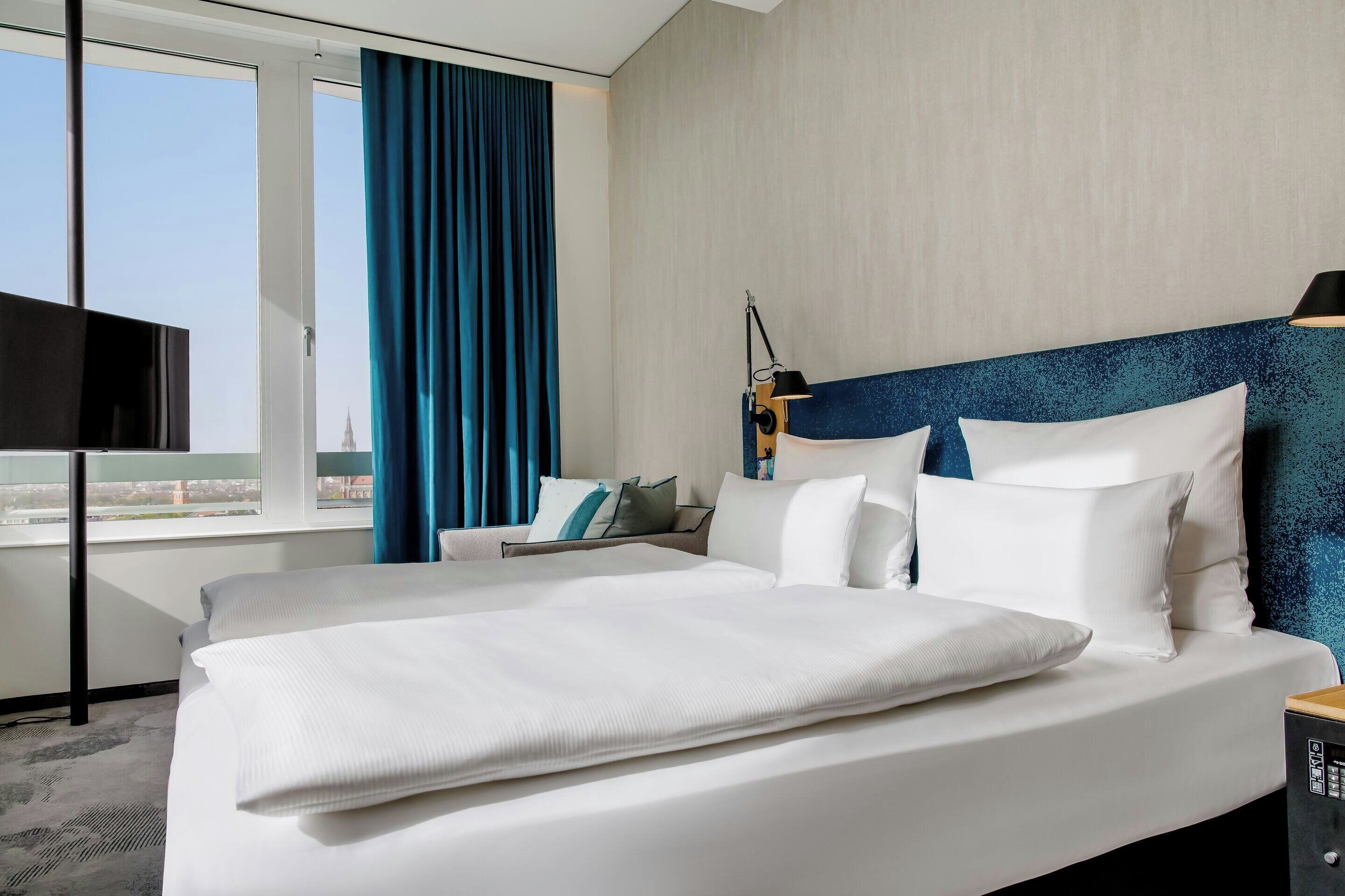 A hotel room with a large bed, a TV, a desk, and a view of the city.