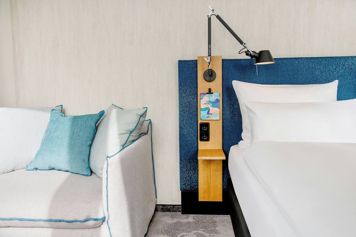 A hotel room with a bed, a sofa, a nightstand with a lamp, and a wall-mounted control panel.