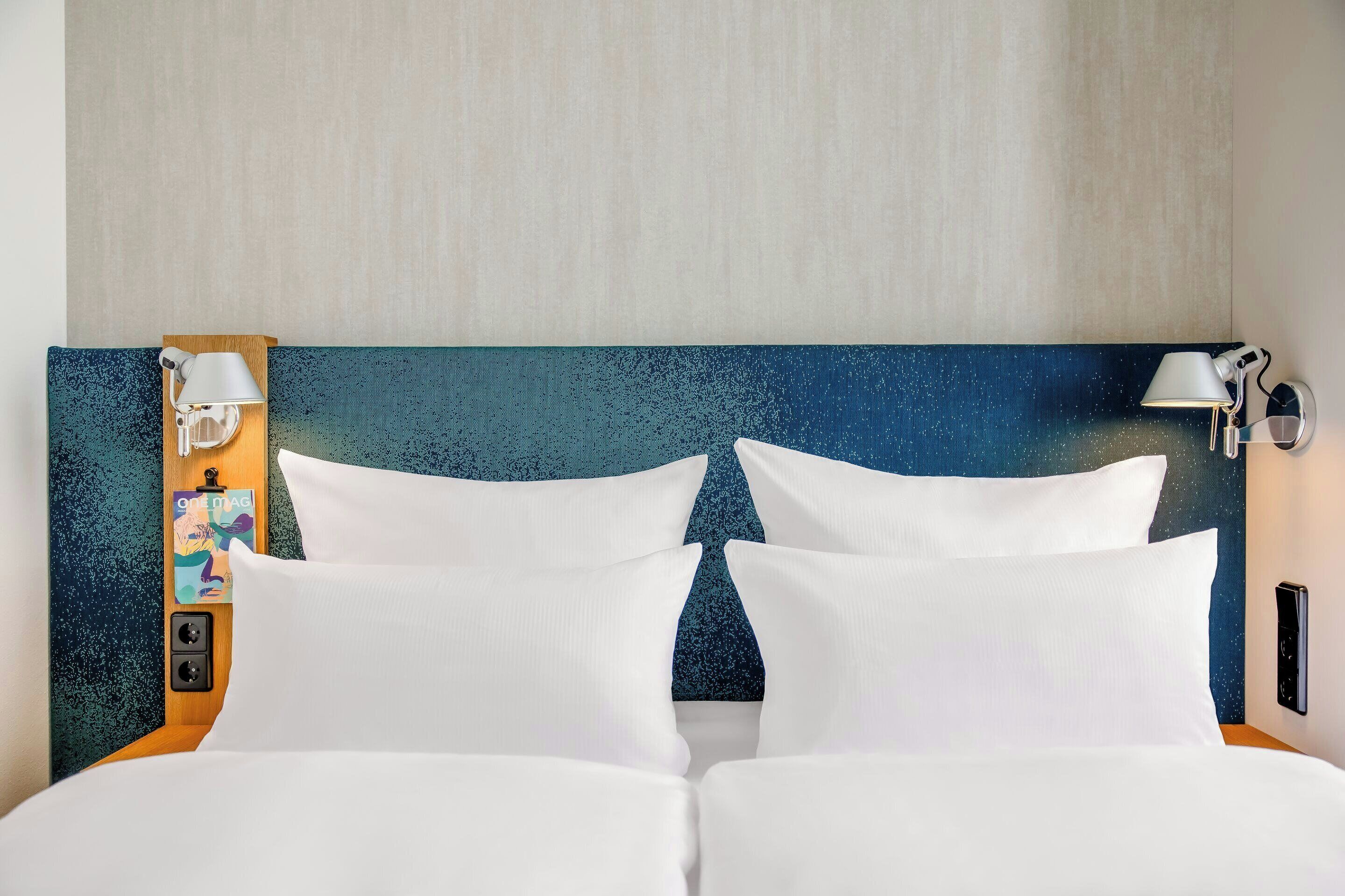A hotel room with a bed, two white pillows, a headboard with a starry night design, and two wall-mounted lamps.
