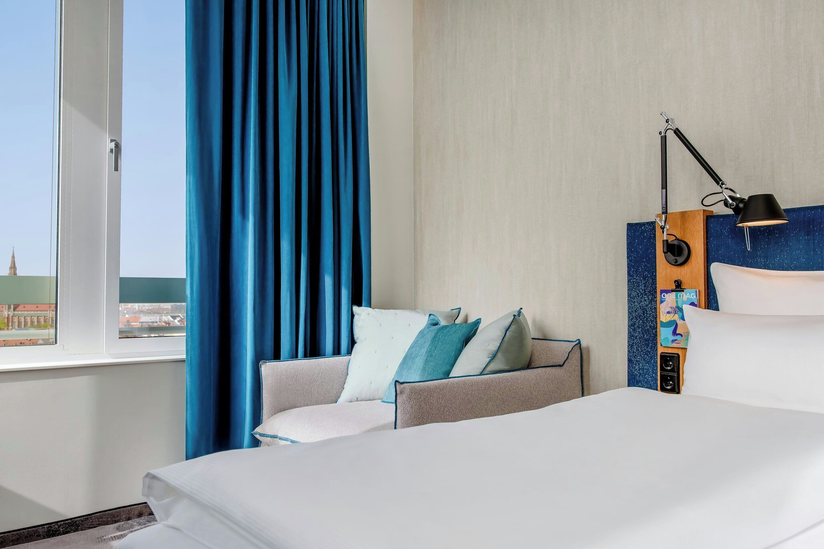 A hotel room with a bed, a seating area with cushions, a window with curtains, and a wall-mounted lamp.