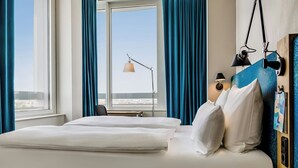 A hotel room with a large bed, a desk, a chair, a desk lamp, and a view of the city. - Motel One München-Campus (Munich)