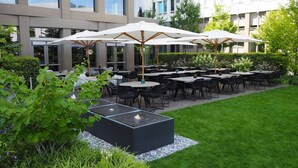An outdoor dining area with multiple tables and chairs, surrounded by greenery and a modern building in the background. - Motel One München-Campus (Munich)