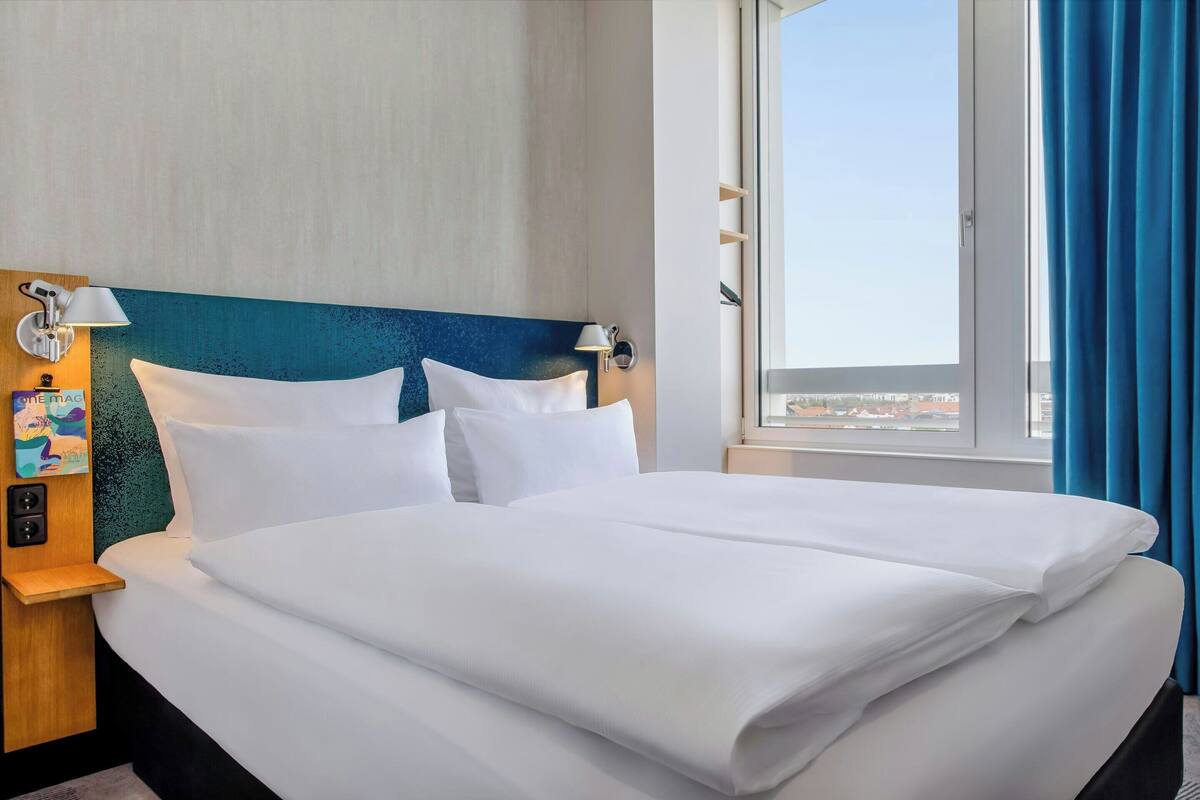 Una habitación de hotel con una cama grande, un cabecero con detalles azules, una mesita de noche con una lámpara y una ventana con cortinas.