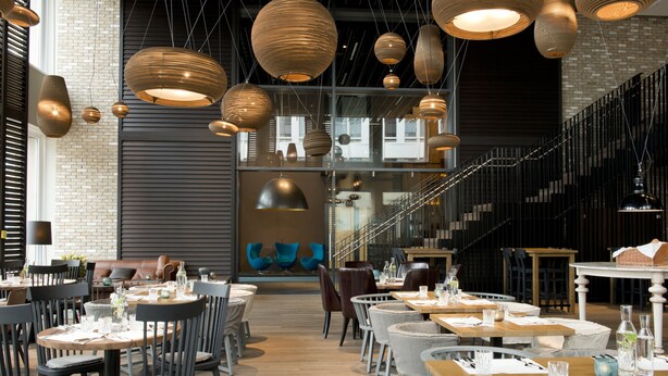 A modern restaurant with multiple dining tables and chairs, large hanging pendant lights, and a staircase leading to an upper level. - Motel One München-Campus (Munich)