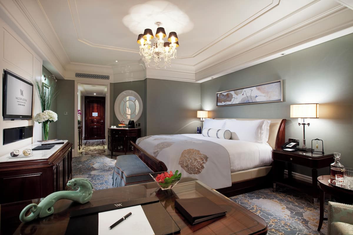 Ein Hotelzimmer mit einem großen Bett, einem Schreibtisch mit Blumenvase, einer Chandelüster und einem Gemälde an der Wand.