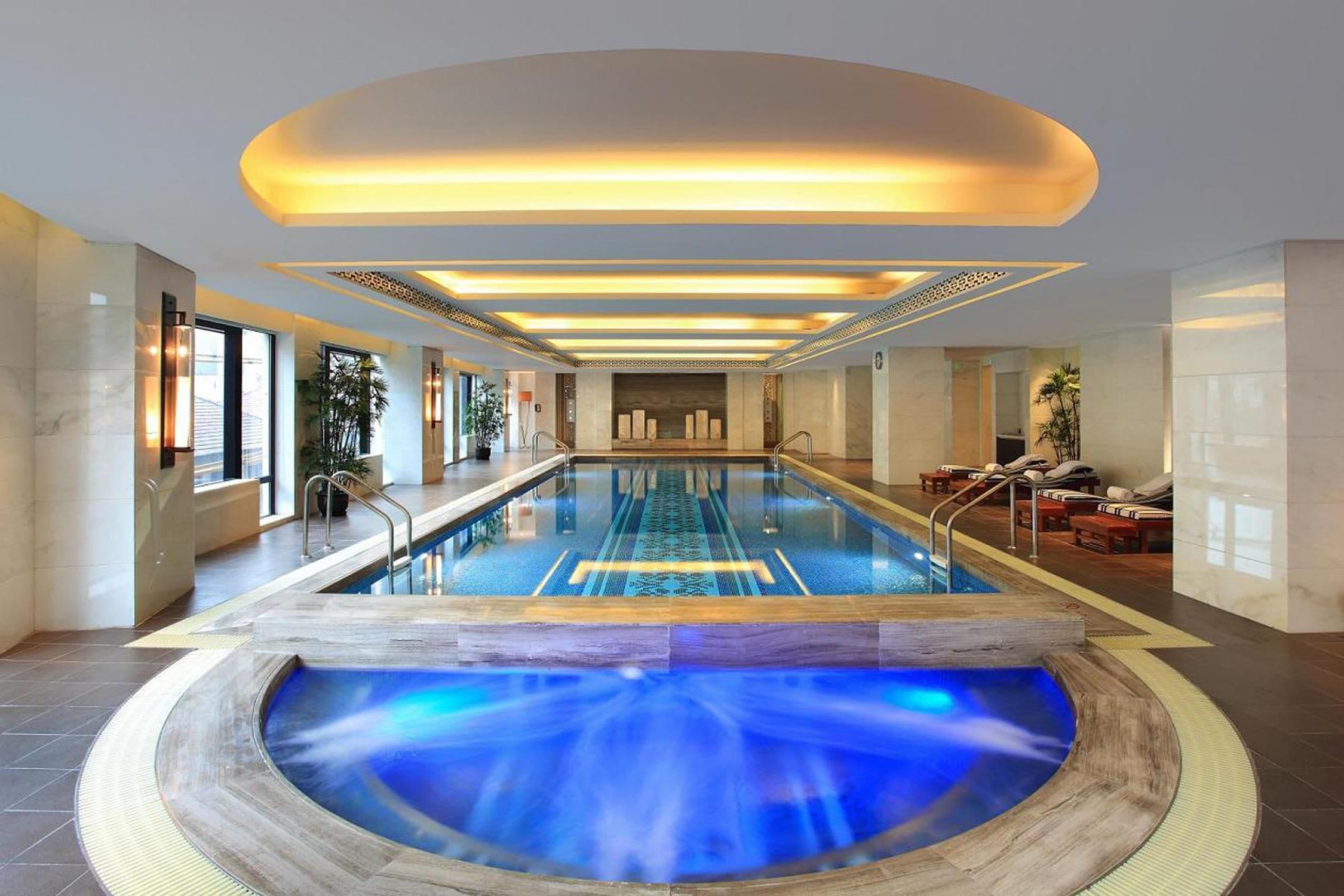A large indoor swimming pool with a central water feature, surrounded by lounge areas and a modern interior design.