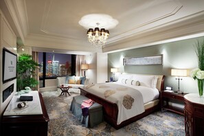 Ein Hotelzimmer mit einem großen Bett, einem Schreibtisch, einem Sessel, einem kleinen Tisch, einer Lampe, einer Vase mit Blumen, einer Pflanze und Blick auf die Stadt.