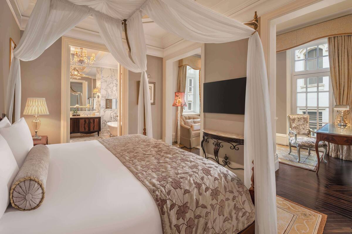 Ein luxuriöses Schlafzimmer mit Baldachinbett, einer Sitzecke mit Fernseher und Blick auf die Stadt.