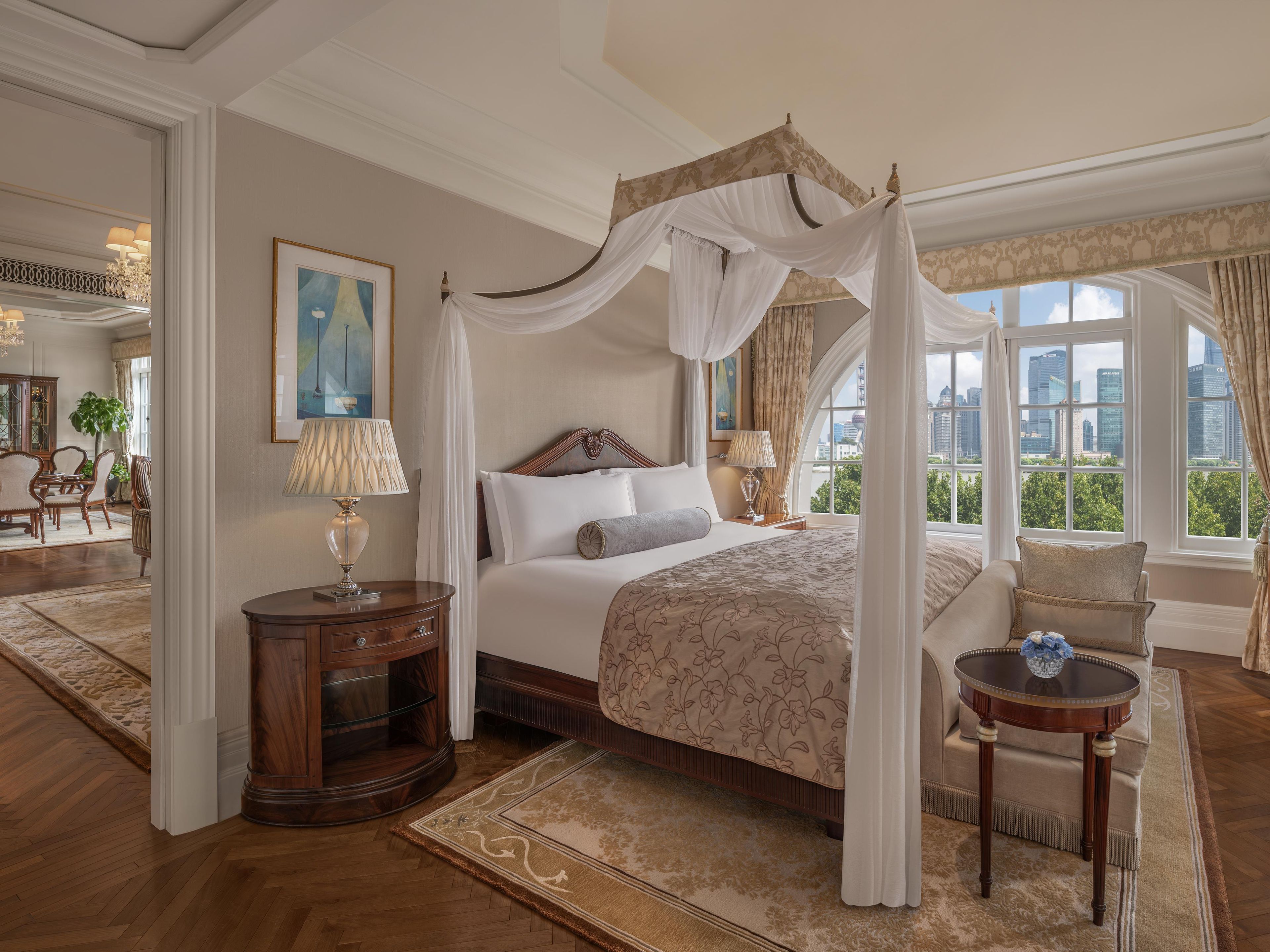 A four-poster canopy bed with a city view, a round wooden nightstand, a small round table with a vase, and a large window with curtains.