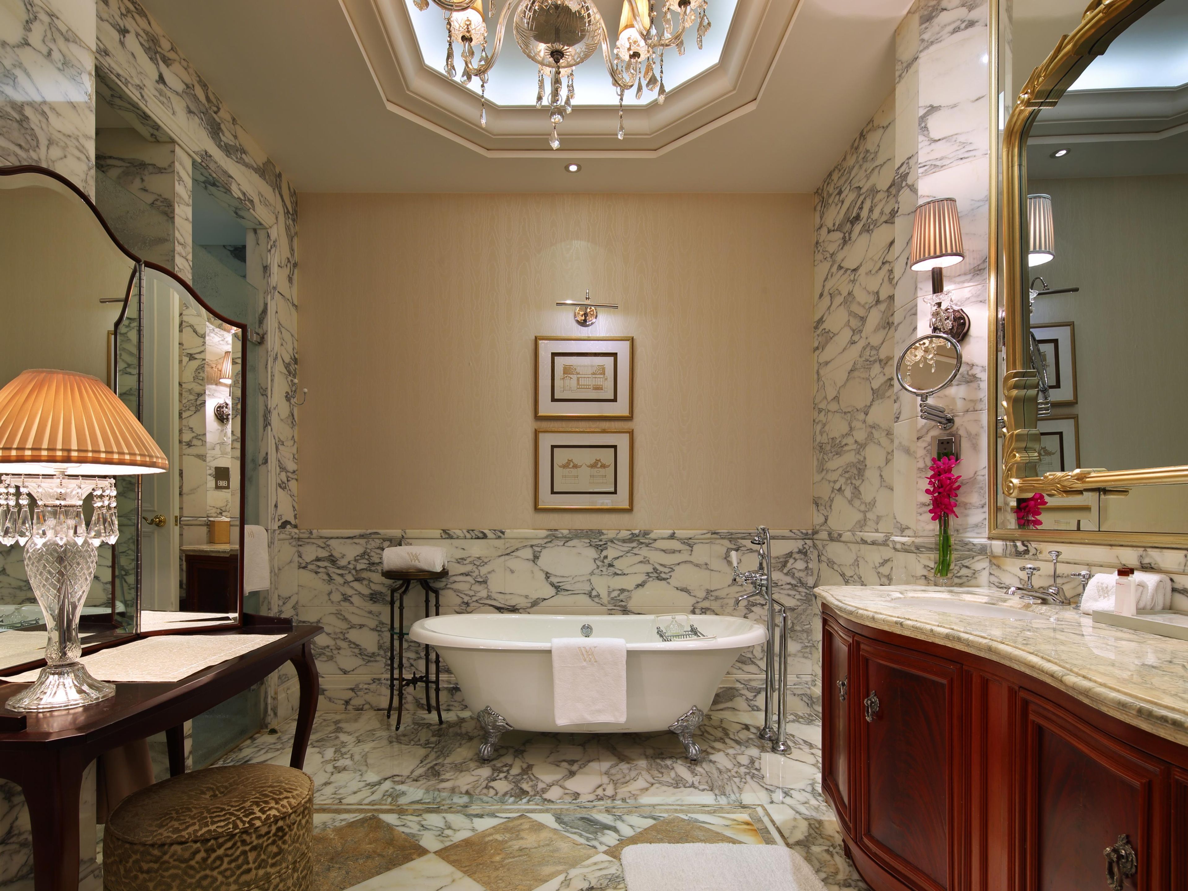 A bathroom with a freestanding bathtub, marble walls, a vanity with a mirror, and a chandelier.