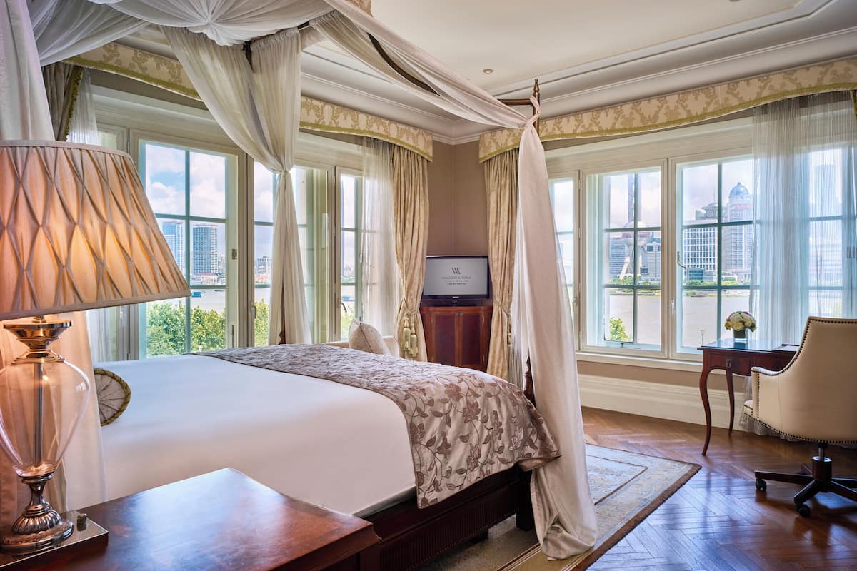 Ein geräumiges Schlafzimmer mit einem großen Bett, einem Schreibtisch und einem Sessel, mit Blick auf die Stadt durch große Fenster.