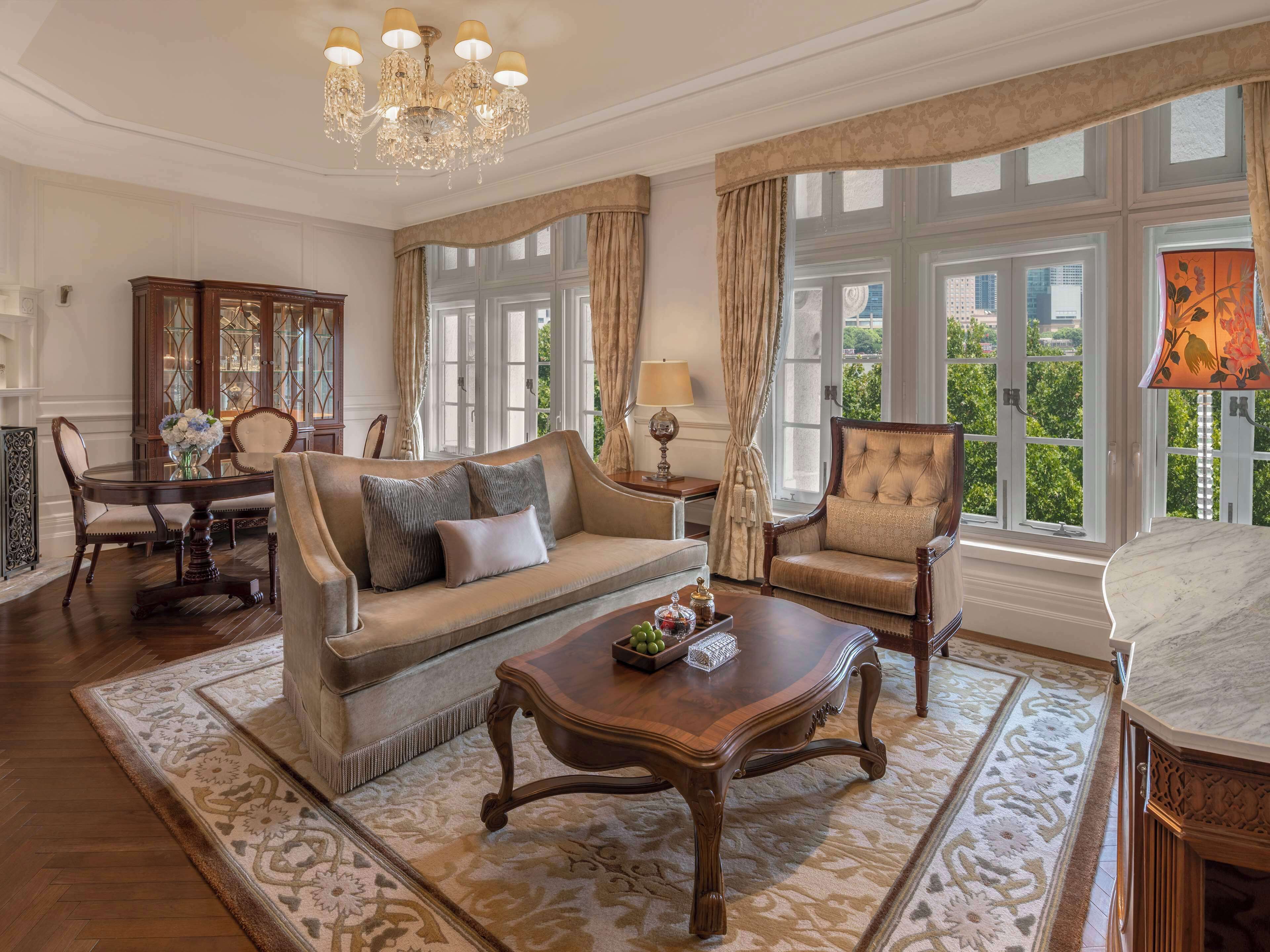 A spacious living room with a sofa, armchair, and coffee table, large windows with curtains, and a chandelier.
