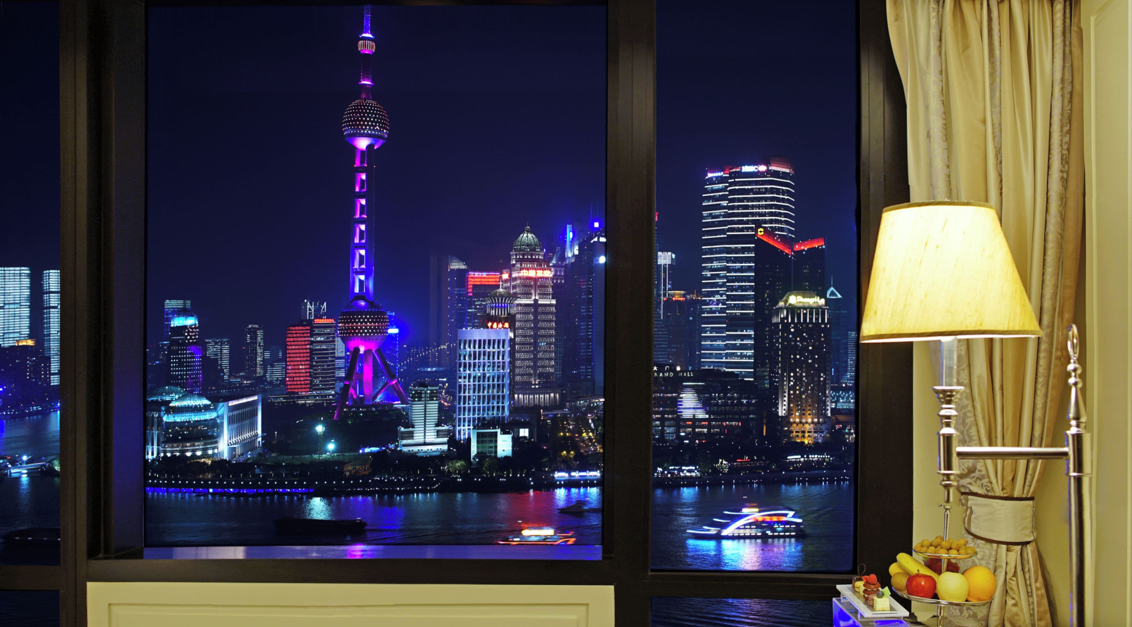 A night view of a cityscape with illuminated buildings and a tower, a window with curtains, and a table with fruits.