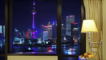 A night view of a cityscape with illuminated buildings and a tower, a window with curtains, and a table with fruits.
