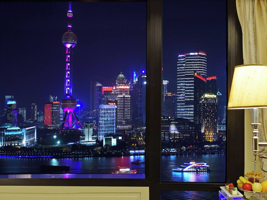 A night view of a cityscape with illuminated buildings and a tower, a window with curtains, and a table with fruits.