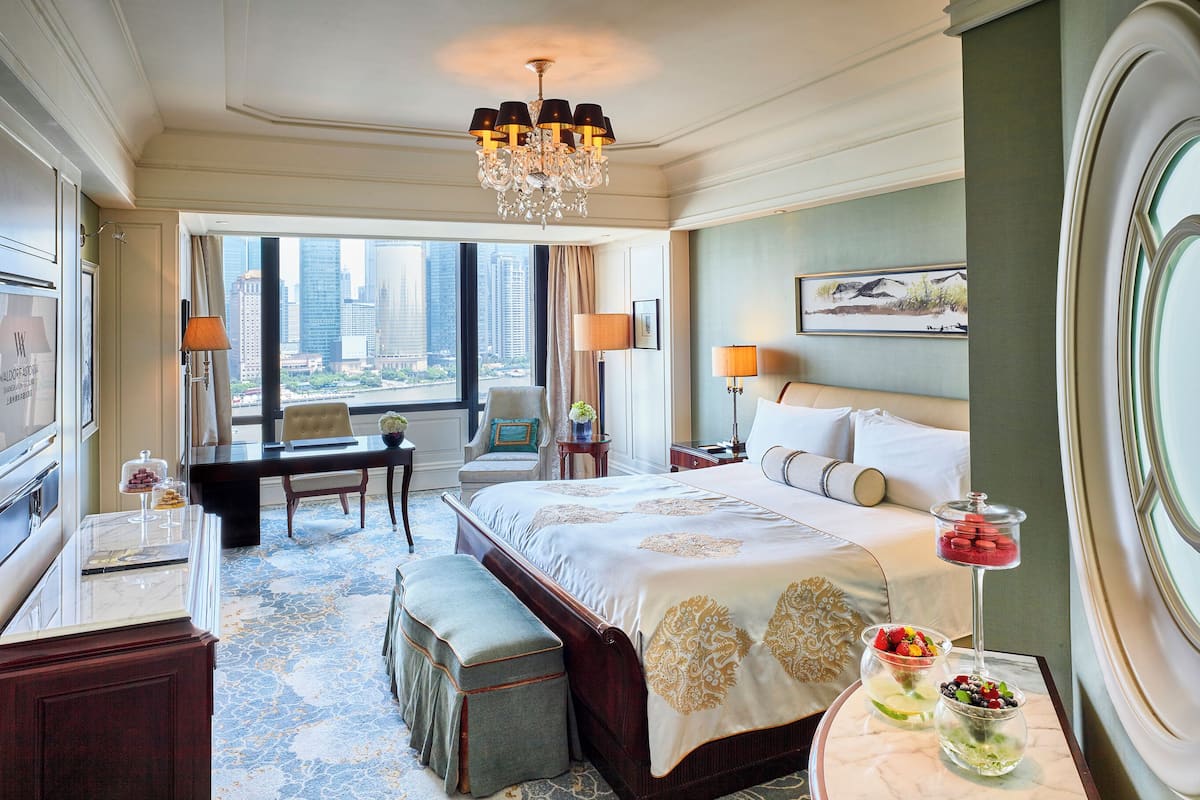 Ein Hotelzimmer mit einem großen Bett, einem Schreibtisch, einem Sessel, einem kleinen Tisch mit Lampe, Blick auf die Stadt und einer Kristallleuchter.