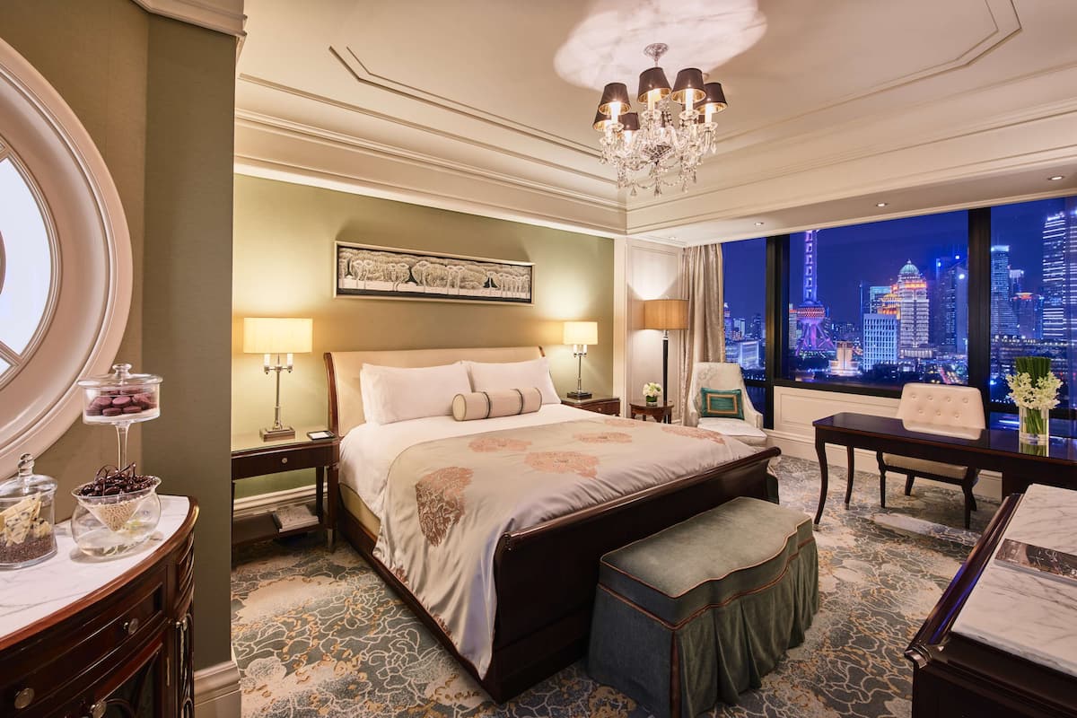 Ein luxuriöses Hotelzimmer mit einem großen Bett, einem Schreibtisch, einem Stuhl und Blick auf die nächtliche Skyline der Stadt.