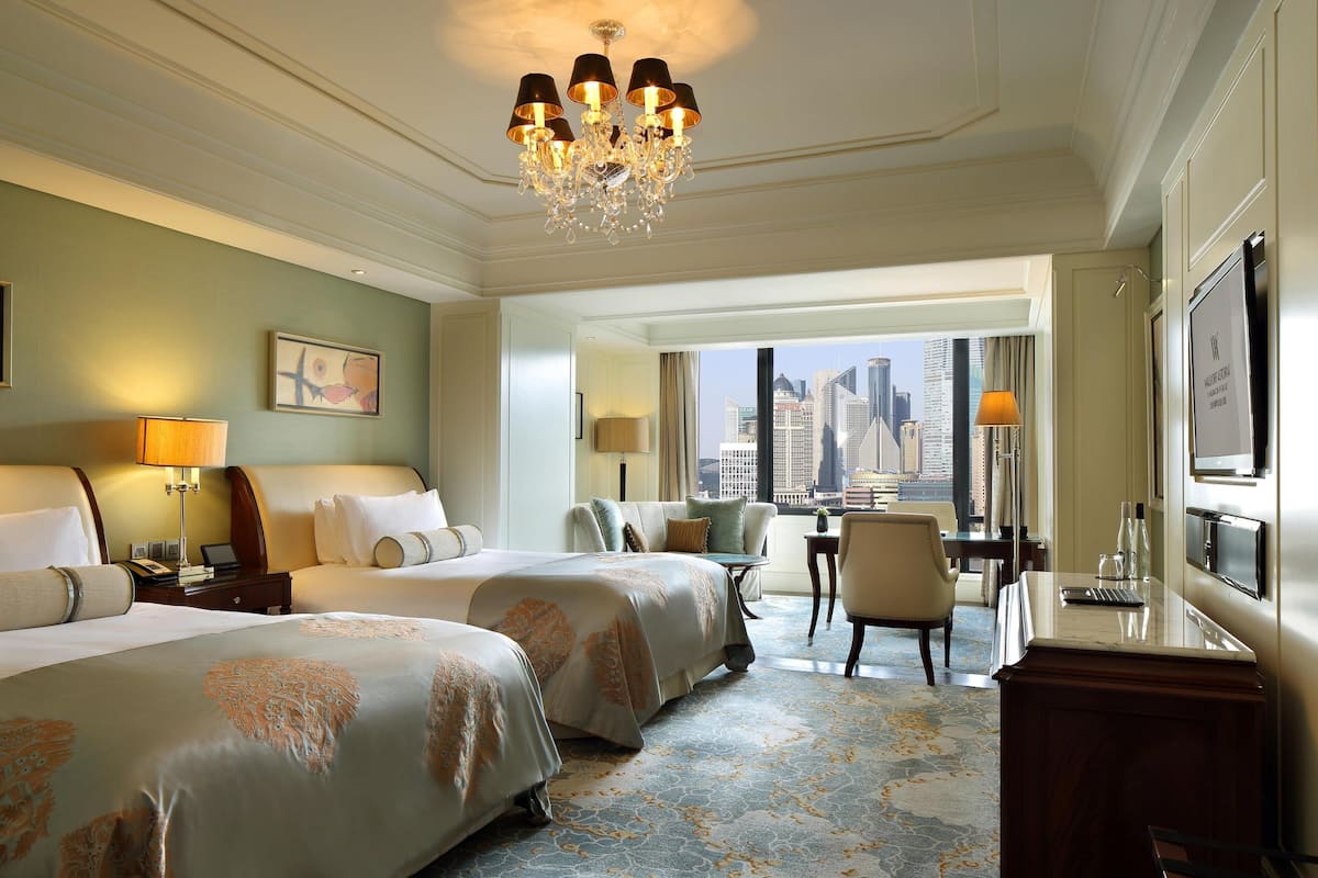 Ein Hotelzimmer mit zwei Betten, einem Schreibtisch, einem Sessel, einem Fernseher und Blick auf die Stadt.