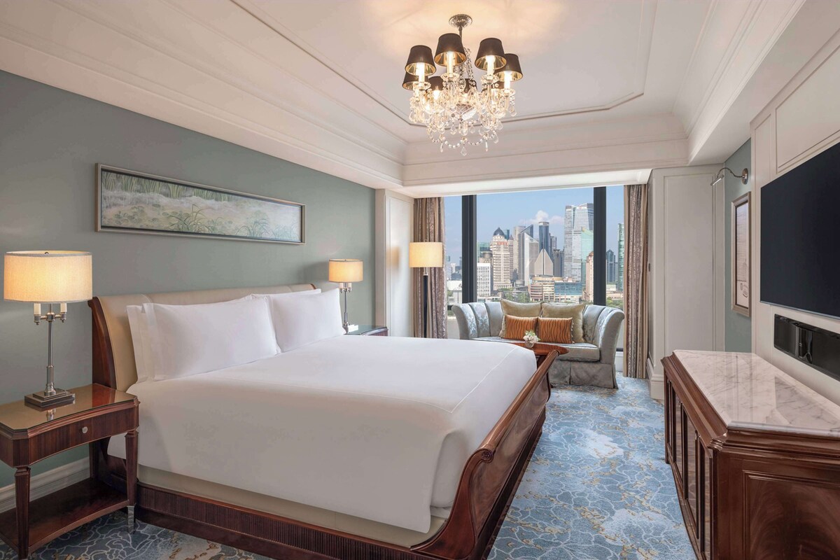 Une chambre d’hôtel avec un grand lit, deux tables de chevet, un lustre, un canapé, une télévision et une vue sur la ville.
