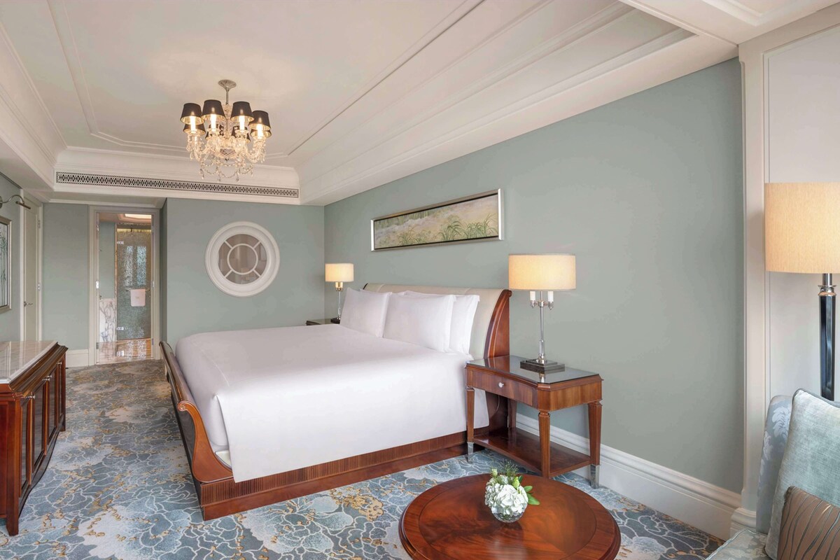 Une chambre d’hôtel avec un grand lit, une table ronde en bois surmontée d’un vase, un lustre et un tableau accroché au mur.