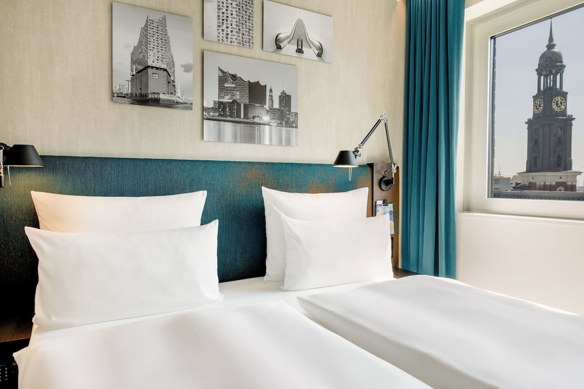 Quarto de hotel com duas camas, cabeceira verde-água e vista para a cidade pela janela.