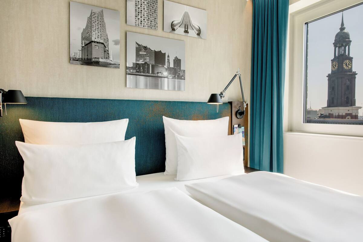 A hotel room with two beds, a teal headboard, and a cityscape view through the window.
