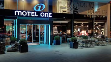 A modern motel with a prominent 'Motel One' sign, outdoor seating area, and a visible interior with a fire feature.