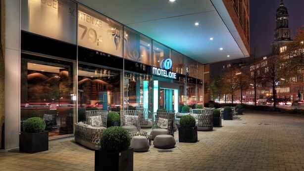 Ein modernes Hotel mit einer gut sichtbaren 'Motel One'-Beschilderung, einem Außenbereich mit Sitzgelegenheiten und einem dahinterliegenden Glockenturm.