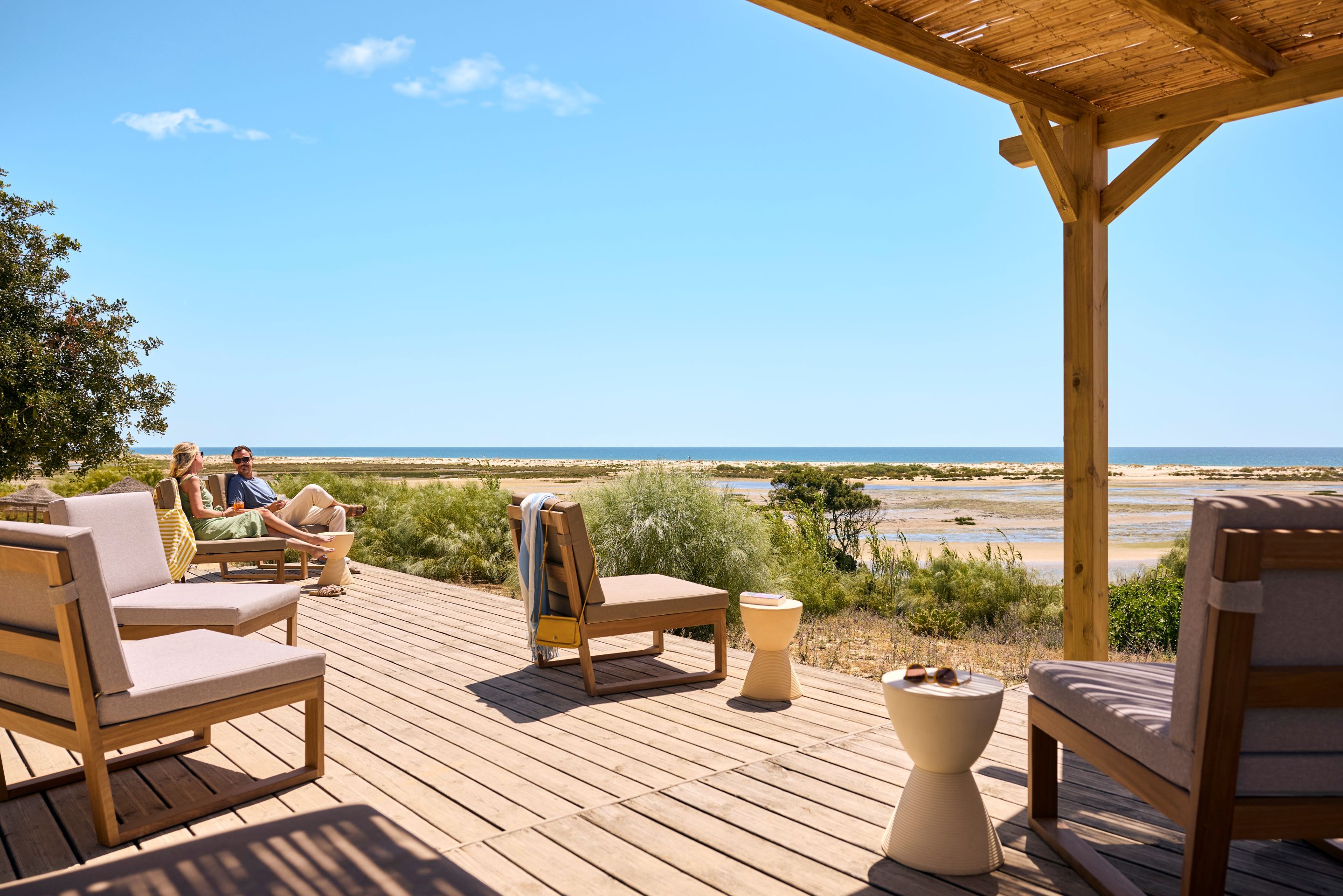 Eine Holzterrasse mit Liegestühlen und einem Tisch, mit Blick auf einen Strand unter blauem Himmel.