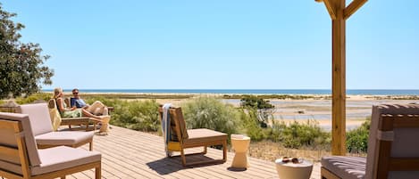 Terraza de madera con sillones de descanso y una mesa, con vista a una playa y un cielo despejado.