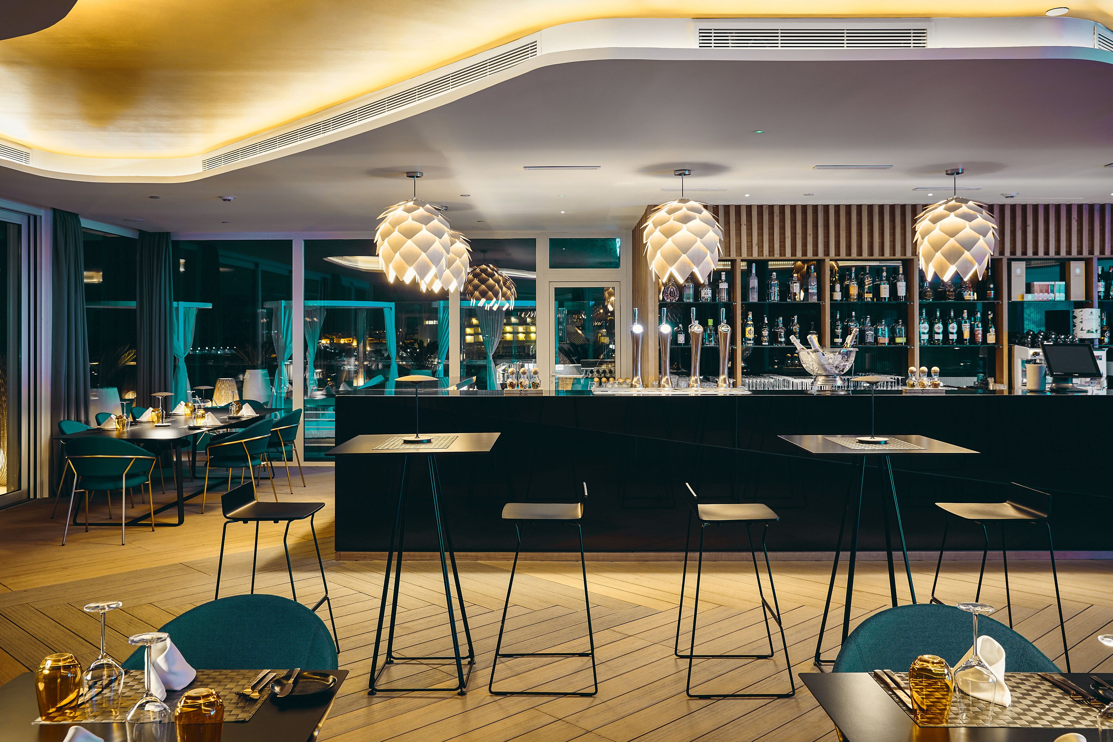 A modern bar area with high stools, a counter with bottles, and a view of the city at night.