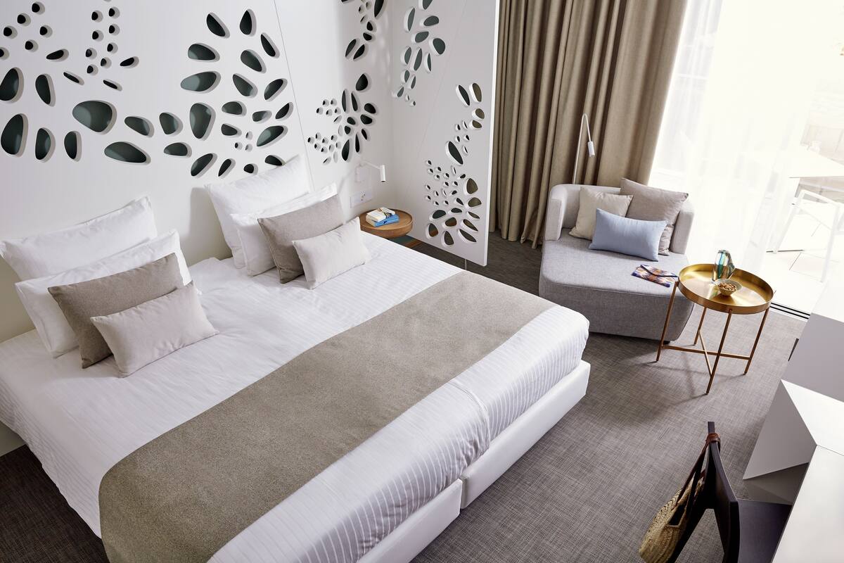 Ett modernt hotellrum med en stor säng, en soffa, ett litet sidobord och en dekorativ väggpanel.