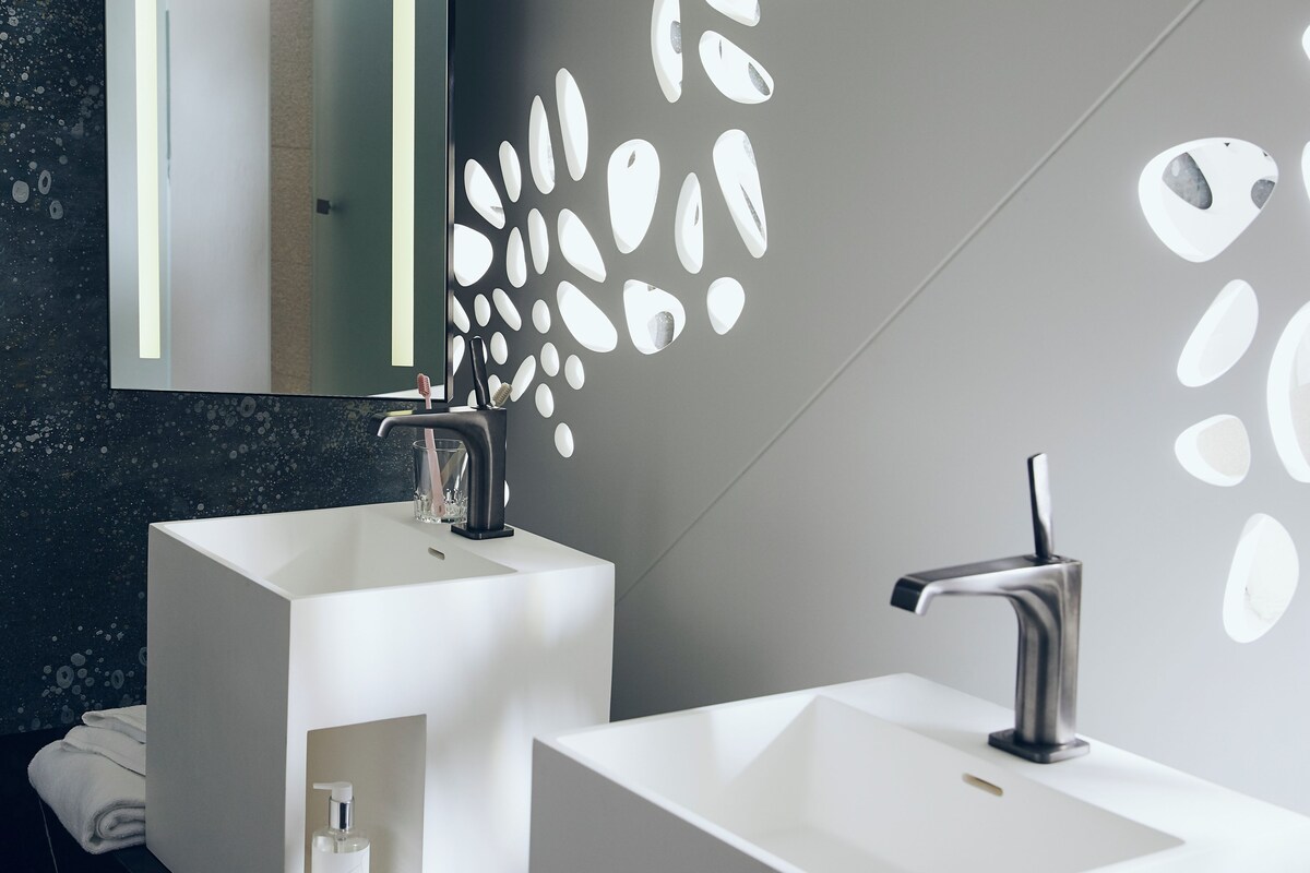 Un baño moderno con dos lavamanos blancos, un espejo grande y una pared con patrones de luz abstractos.