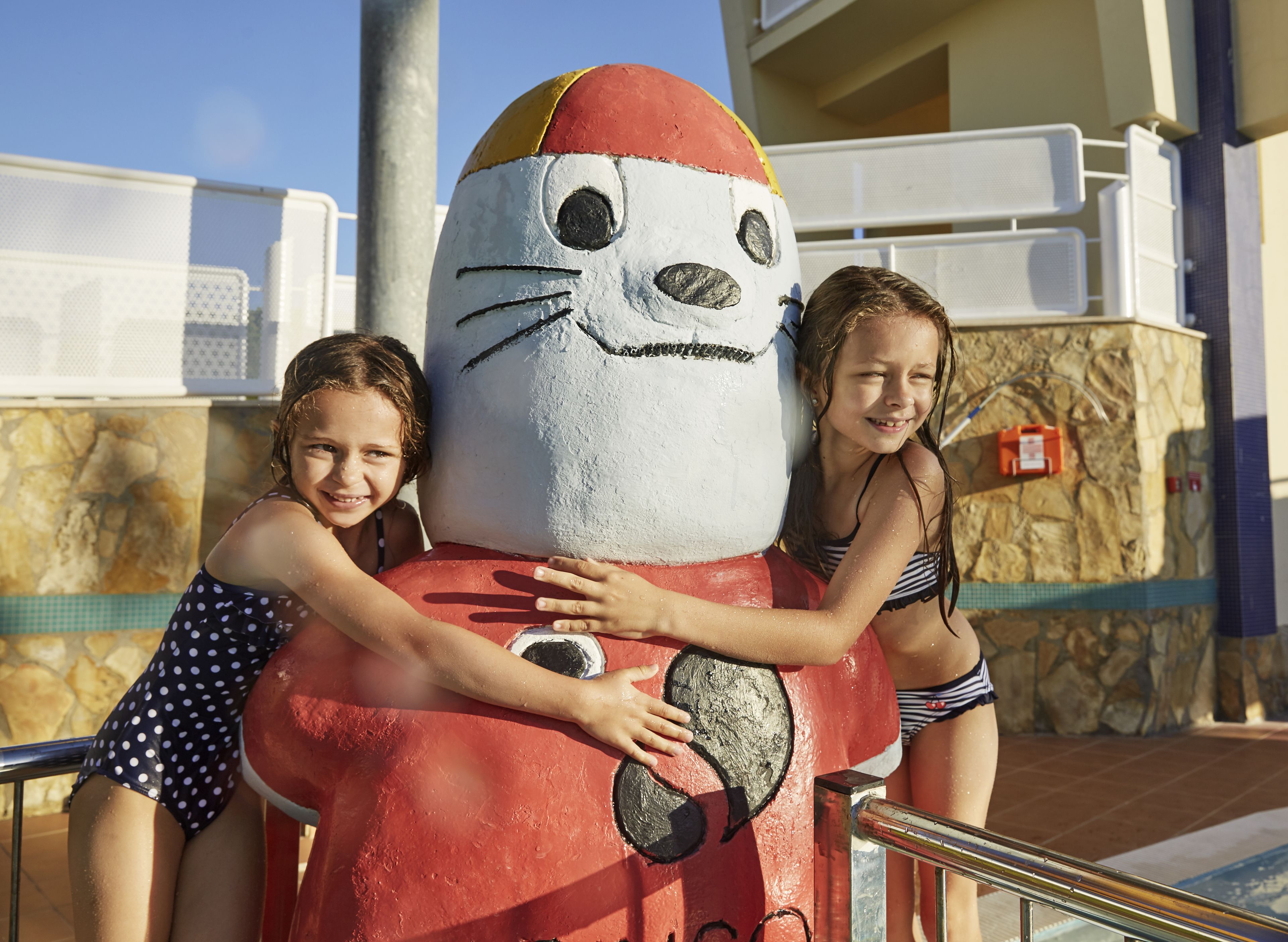 A large mascot sculpture with a smiling face and a red body, located near a poolside area with stone walls and a metal railing.
