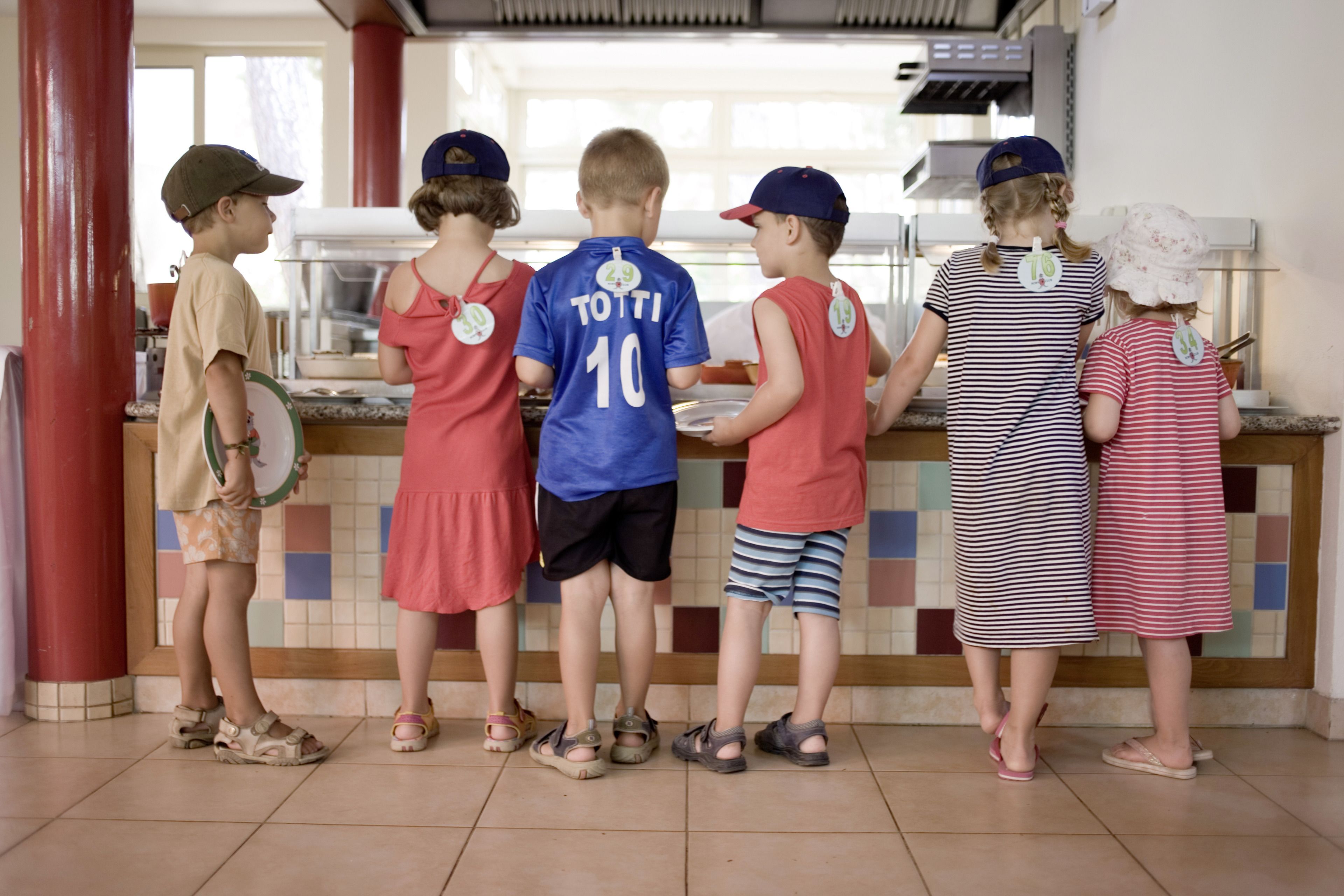 A group of children standing in line at a counter, possibly in a cafeteria or food service area.