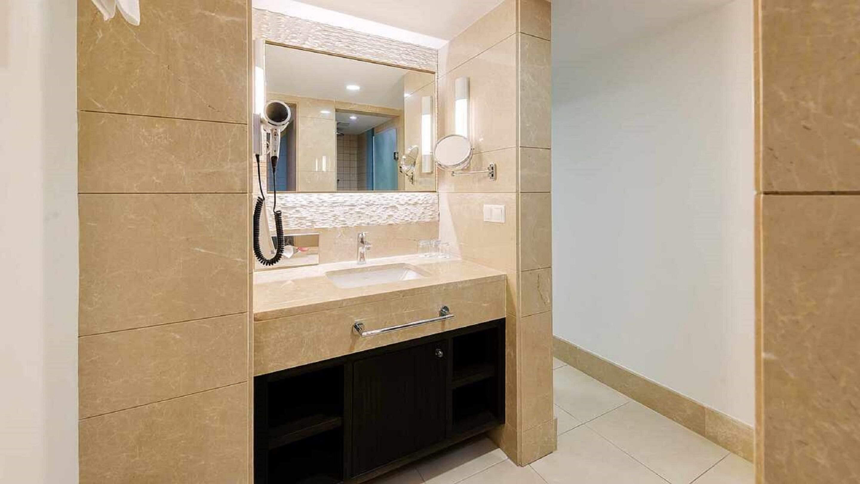 A bathroom with a beige tiled wall, a sink, a mirror, a hairdryer, and a small cabinet.
