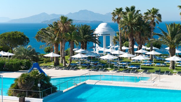 A pool area with lounge chairs and umbrellas, a water slide, and a view of the sea and mountains. - ROBINSON DAIDALOS (Kos)