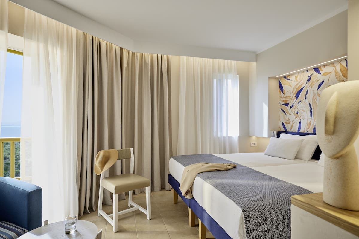 A hotel room with a bed, a chair, a small table, and a view of the outdoors.