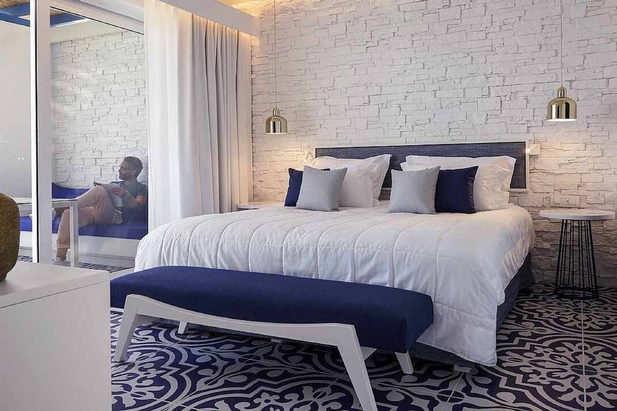 A modern bedroom with a white brick wall, a large bed with multiple pillows, a bedside table, and a small round table with a lamp.
