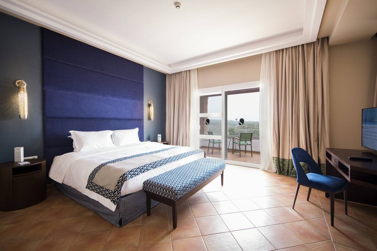 Quarto de hotel com uma cama grande, uma escrivaninha, uma cadeira e varanda com vista.