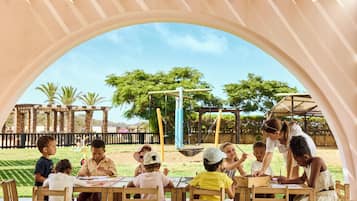 Children learn and play under a shaded area with tables and a playground in the background.