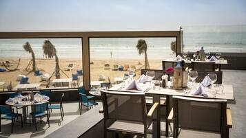 An outdoor dining area with tables and chairs, overlooking a beach with palm trees and the ocean.