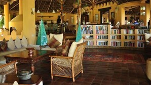 A spacious lounge area with a high wooden ceiling, a central seating area with a wicker chair and a table, and a bookshelf filled with books. - Kinasi Lodge (Mafia Island)