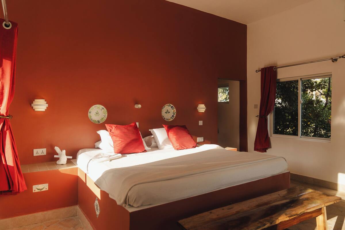A bedroom with a large bed, red curtains, a wooden bench, and a window with a view of trees.